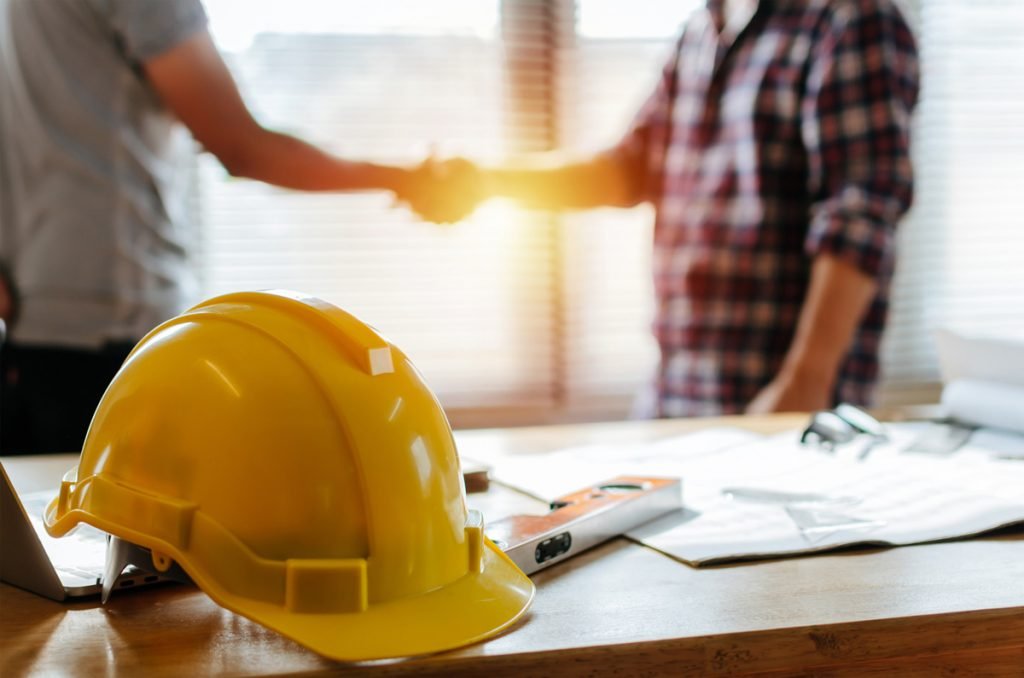 Blog 25 Two people shaking hands behind a yellow hardhat on a desk in El Paso.